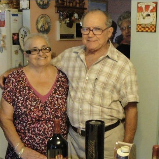 An elderly couple stands together in a warm kitchen, smiling happily during a family gathering.