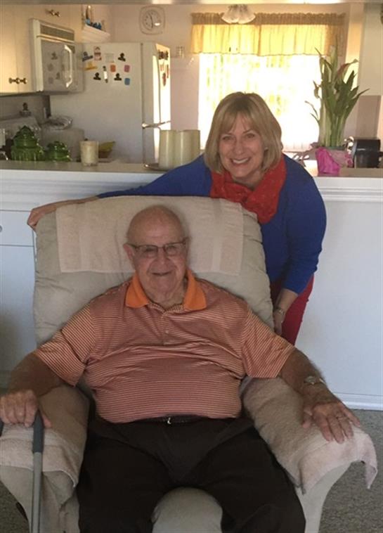 A caregiver and an elderly man share smiles in a well-lit, cozy living room.