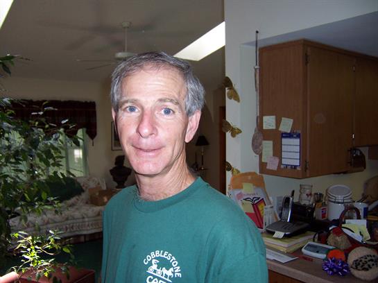 A man with gray hair is smiling in a sunlit room, surrounded by plants and home decor.