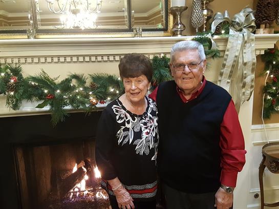 An elderly couple joyfully stands by a decorated fireplace in a cozy living room.