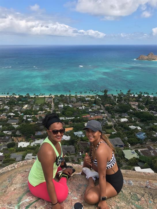 Two friends smile while taking in stunning coastal views from a mountain peak on a sunny day.