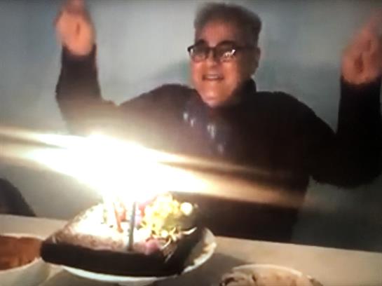 A person joyfully celebrates a birthday with a cake adorned with glowing candles in a dim setting.