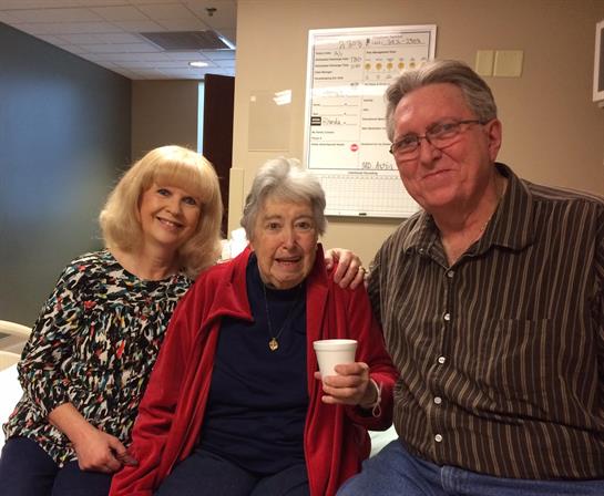 Two family members smile alongside an elderly woman, enjoying a warm moment during a visit.