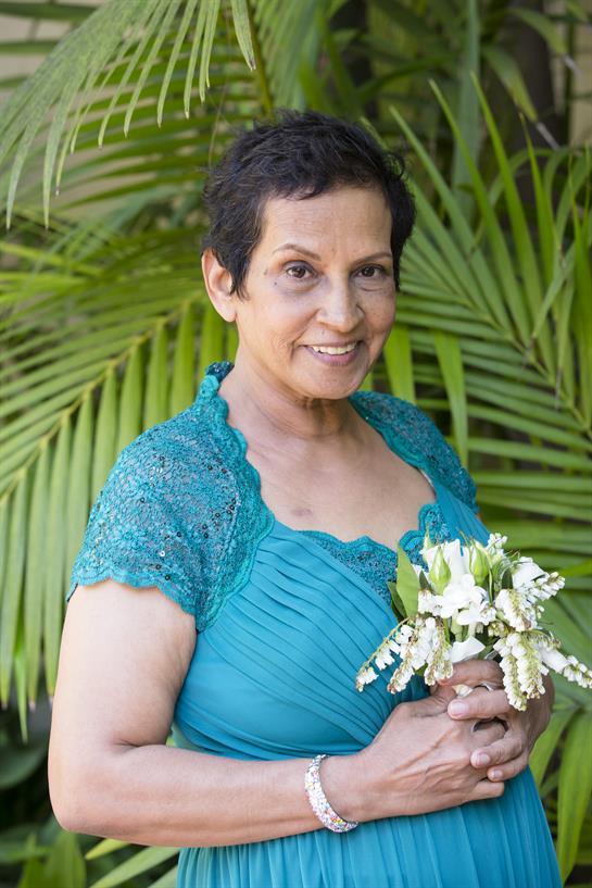A woman stands proudly in a blue dress, holding a bouquet of white flowers amidst lush greenery.