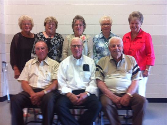 Nine seniors sit together, smiling warmly, sharing memories at a community hall event.