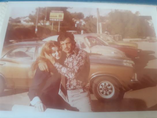 A man and woman embrace happily outdoors near a vintage vehicle on a sunny day in the 1970s.