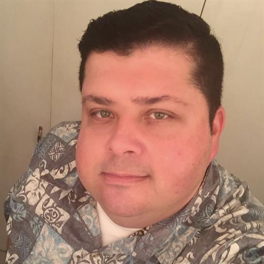 A man with short dark hair and green eyes is smiling while wearing a floral-patterned shirt.