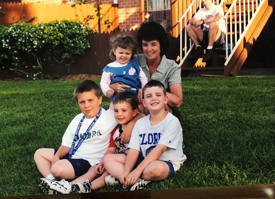 Five kids sit on the grass as an adult holds a smiling toddler, enjoying their time.
