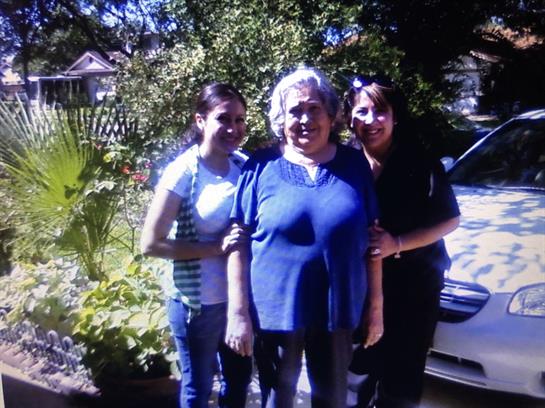 Three women smile together in a lush garden, surrounded by greenery and flowers, on a bright day.