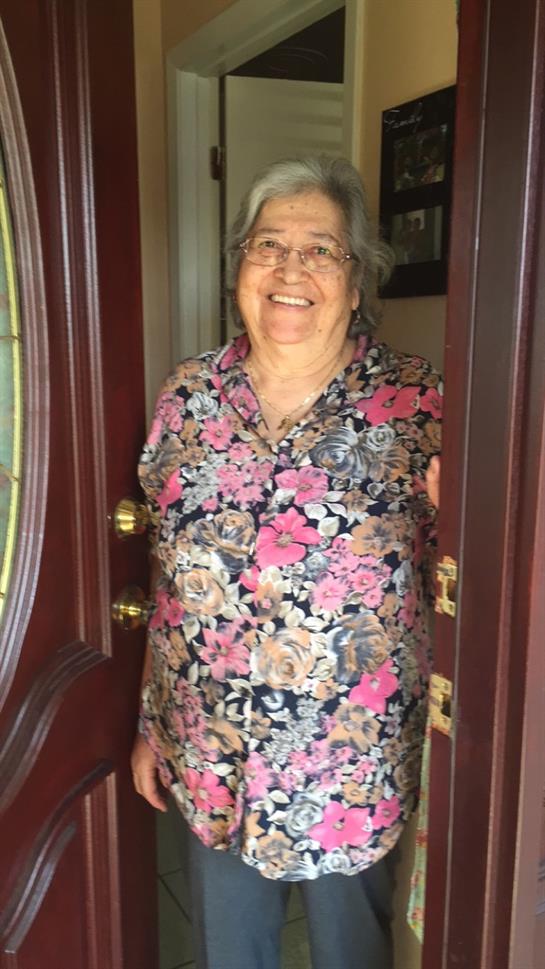 An older woman stands smiling at her front door, welcoming guests with warmth and joy.