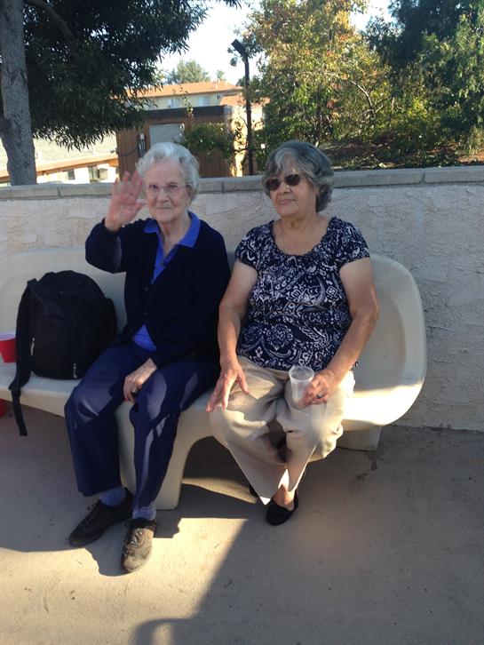 Two senior women sit together on a bench, smiling and greeting passersby in a sunny park.