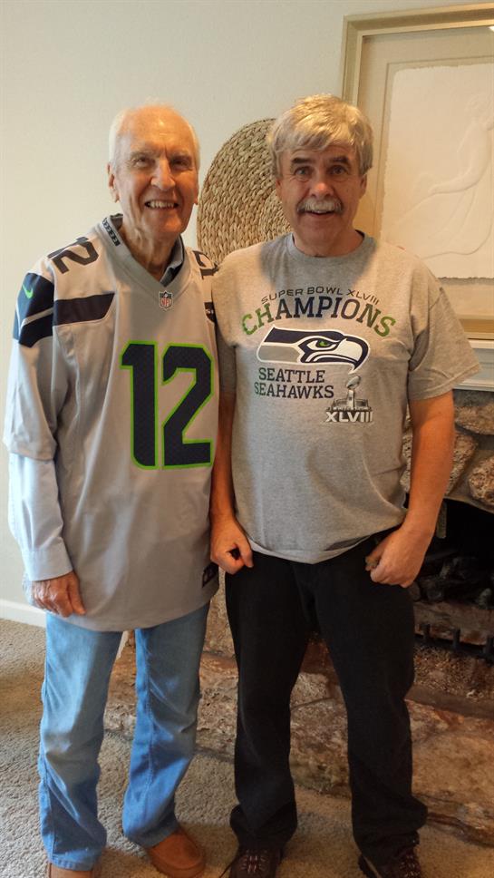 Two men smile while wearing Seattle Seahawks jerseys, celebrating their favorite team.
