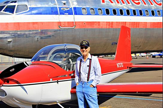 A pilot in uniform poses beside a small red aircraft with a retro airliner behind at an airport.