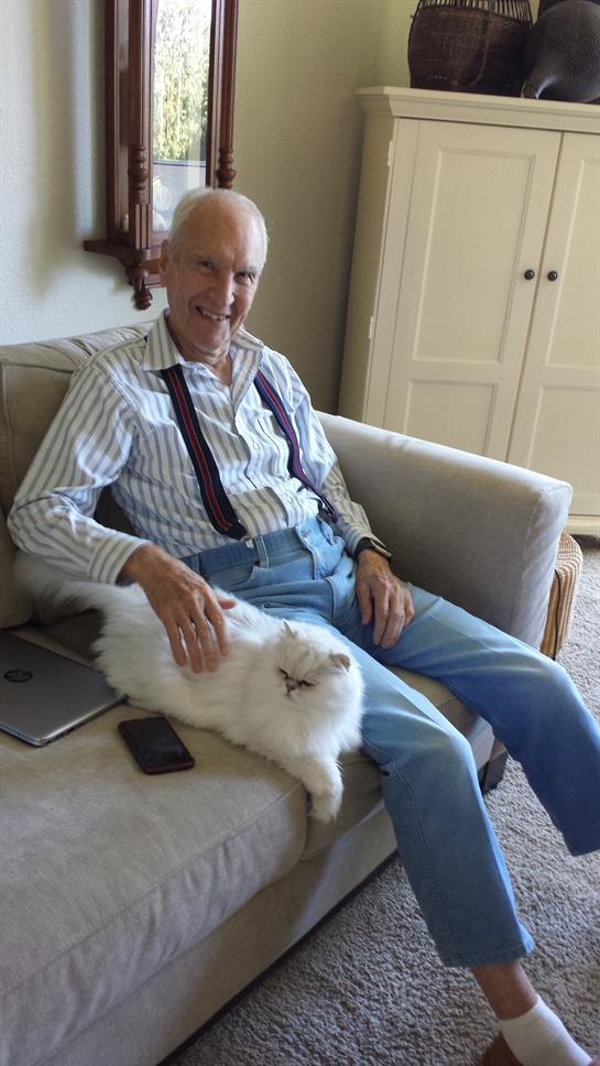 Older gentleman relaxes on a couch while petting a fluffy white cat in a cozy living room.