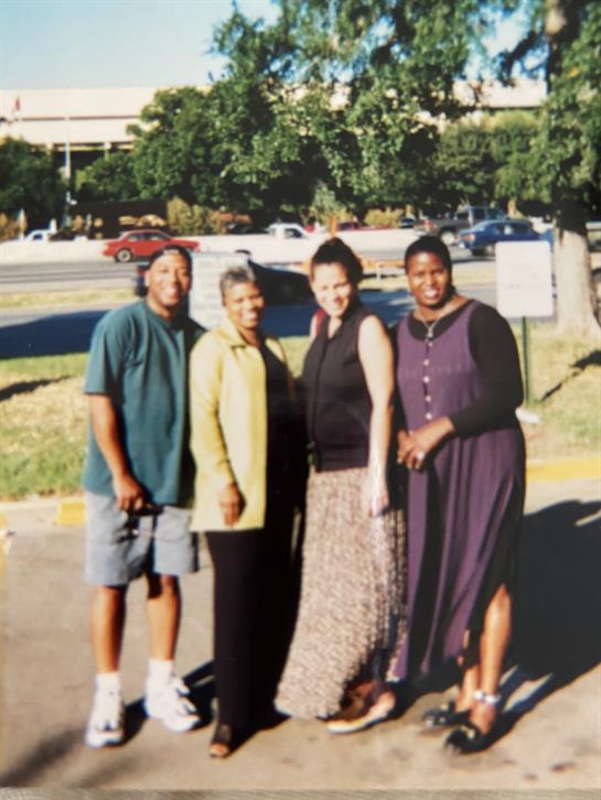 Four friends stand together smiling outdoors, enjoying a sunny day near a busy street.