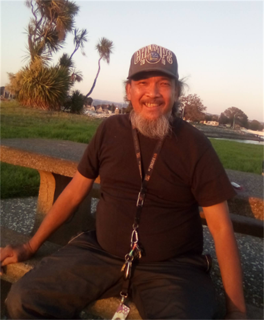 A man laughs while sitting on a bench in a park, enjoying a sunset at the waterfront.