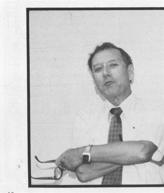 A man dressed in formal clothing stands thoughtfully, leaning against the wall with glasses in hand.