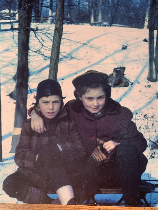 Two children sit closely together on the snow, enjoying winter in a forested area.