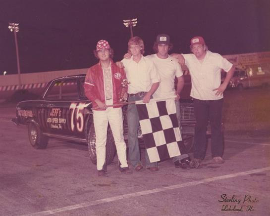 Friends pose together with a classic race car after winning at the track during the evening.