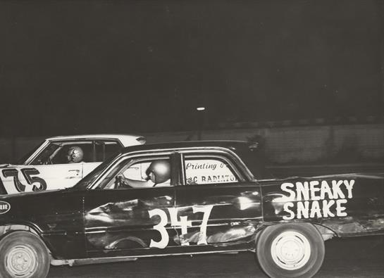 Two race cars drive side by side under bright lights at a vibrant dirt track late in the evening.