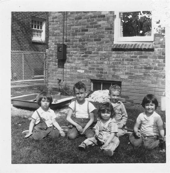 Five children sit on the grass near a brick house, enjoying a sunny day outdoors.