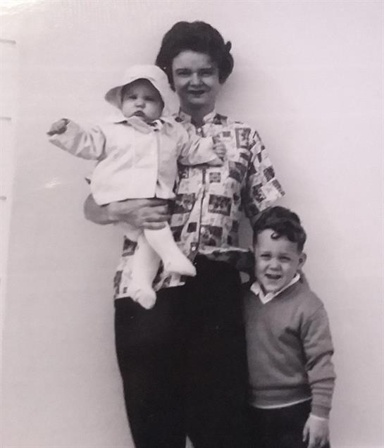 A woman holds a baby while a young boy stands beside her, all smiling against a white background.