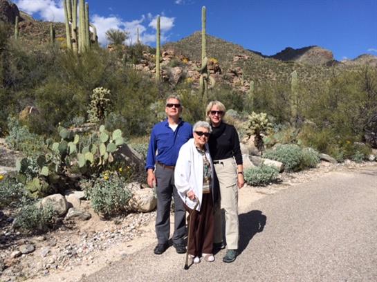 Three people pose together in a sunny desert setting surrounded by cacti and mountains.