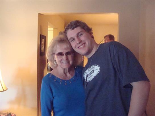 A young man stands next to an elderly woman in a warm, inviting room, both smiling joyfully.