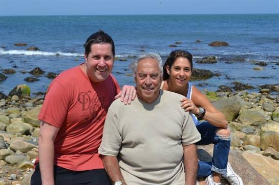 A family poses happily by the ocean, enjoying a sunny day, with waves lapping at the shore.