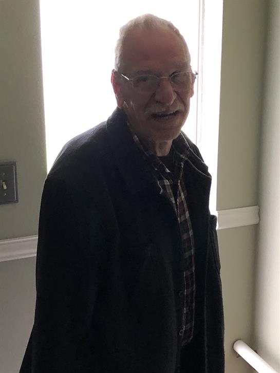 An elderly man stands in a well-lit room, wearing glasses and smiling cheerfully at the viewer.