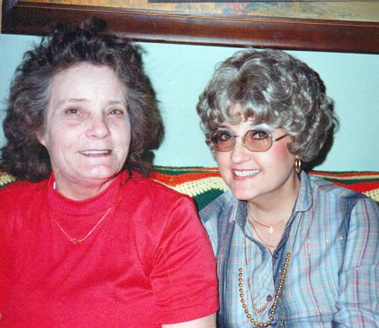 Two women sit closely on a sofa, smiling with enjoyment during a relaxed gathering at home.