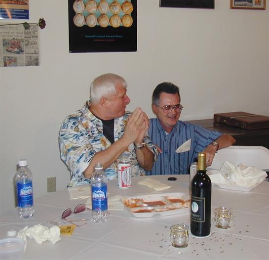 Two men engage in lively conversation at a gathering, surrounded by food and drinks on a table.