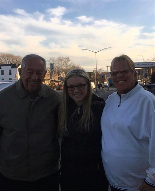 Three individuals stand together in a city setting, smiling happily during a sunny afternoon.
