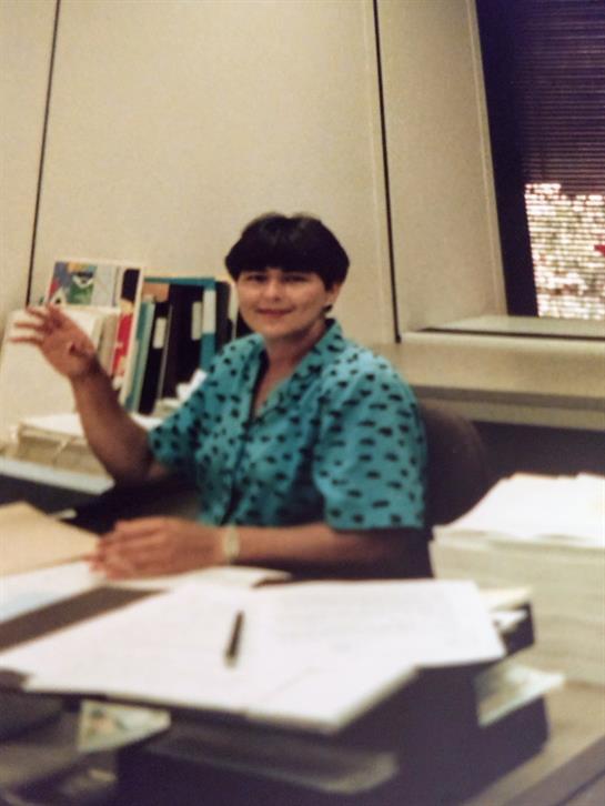 A woman with short hair smiles while seated at her desk, surrounded by documents and books.