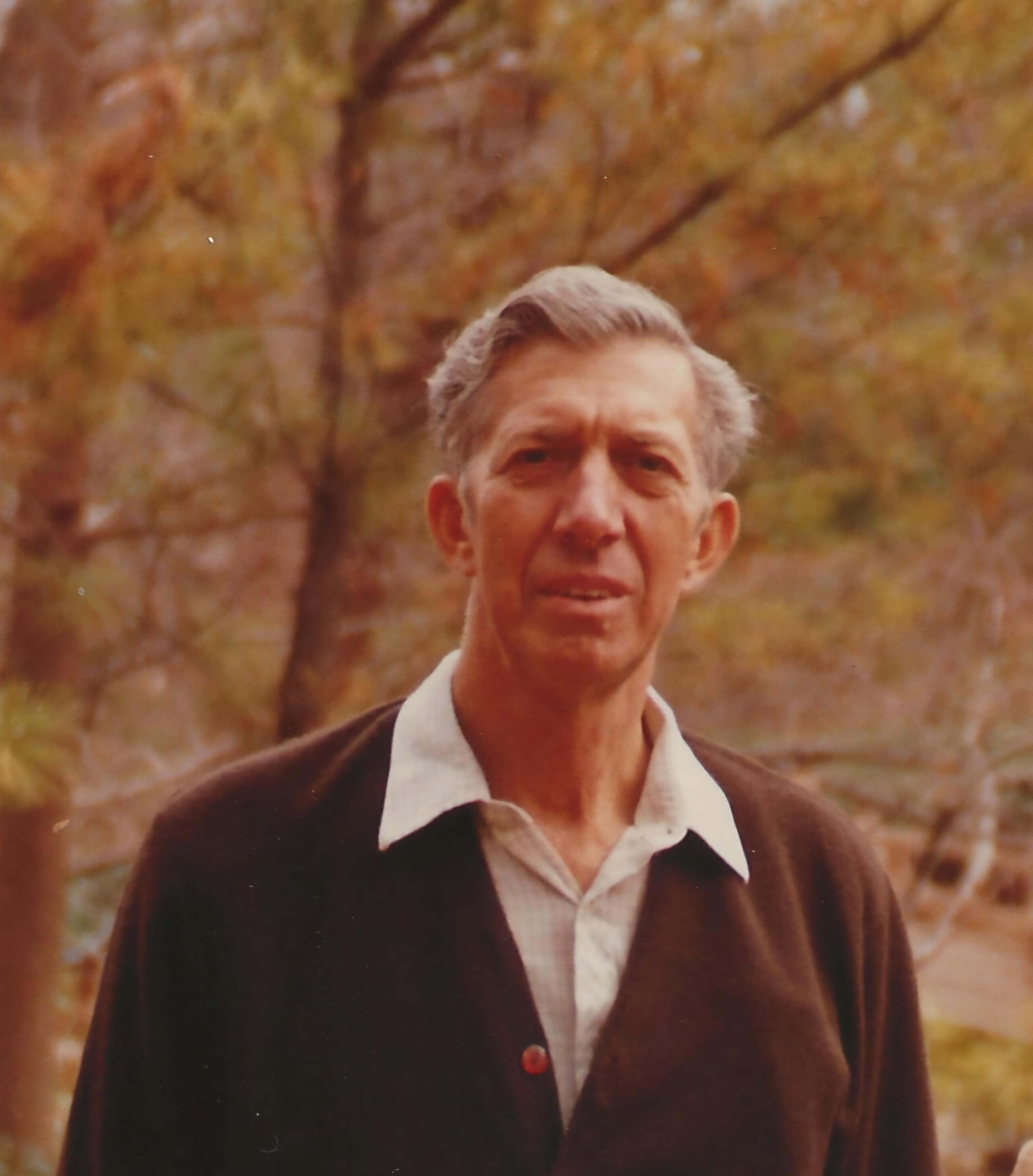 Elderly man poses outdoors surrounded by lush trees on a sunny day, looking calm and content.