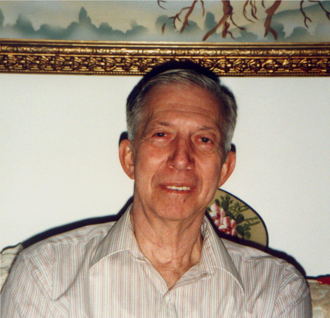 An elderly man, dressed casually, smiles while sitting against a decorated wall.