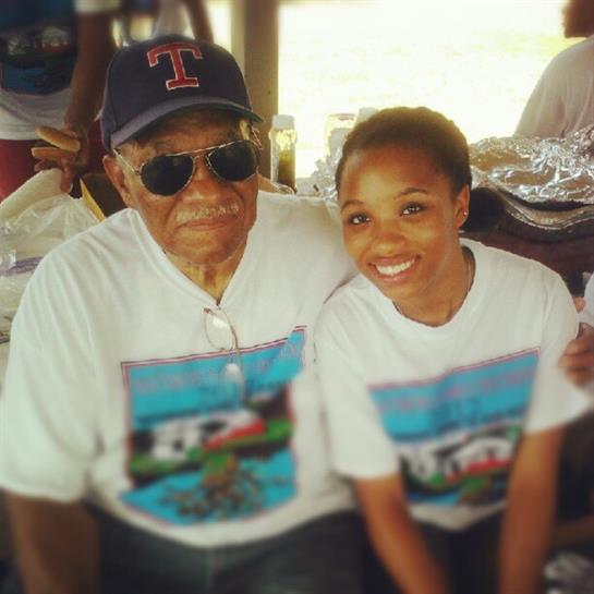 An elderly man wearing sunglasses and a cap sits with a young woman at a sunny outdoor event.