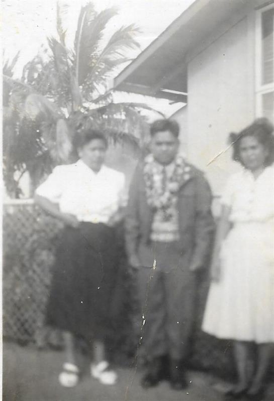 Three individuals stand together near a house surrounded by tropical plants on a sunny day.