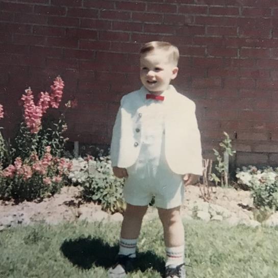 A young boy is smiling while wearing a white suit with a bow tie, surrounded by colorful flowers.