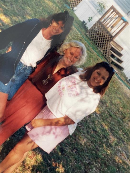 Three women smile and pose in a grassy area, enjoying a sunny day together outdoors.