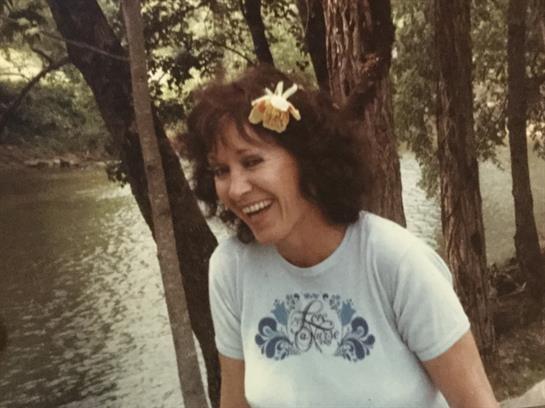 Woman smiles while sitting by a river, surrounded by trees and enjoying a sunny atmosphere.