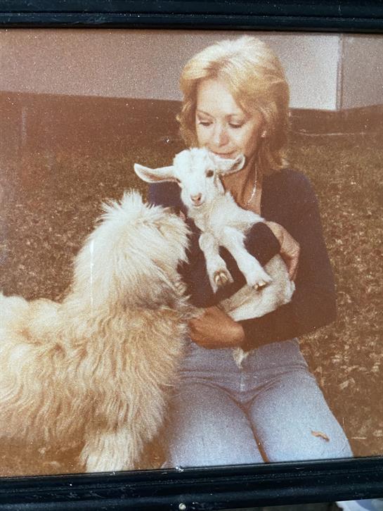 A woman gently cradles a baby goat in her arms as a fluffy dog approaches, both in a grassy setting.