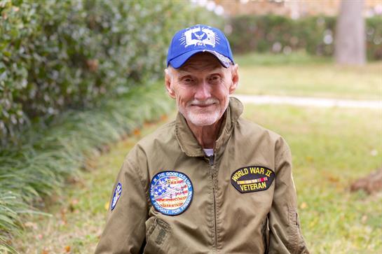 A smiling elderly veteran sits in a park wearing a military jacket and cap, enjoying the day.