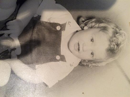 A young child wearing overalls and a white shirt sits on the floor, smiling and looking curious.