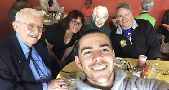 Five people smile for a selfie while enjoying a lively lunch together at a restaurant.