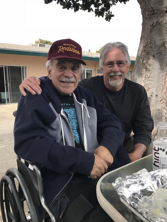 Two older men are smiling and enjoying each other's company at an outdoor table.