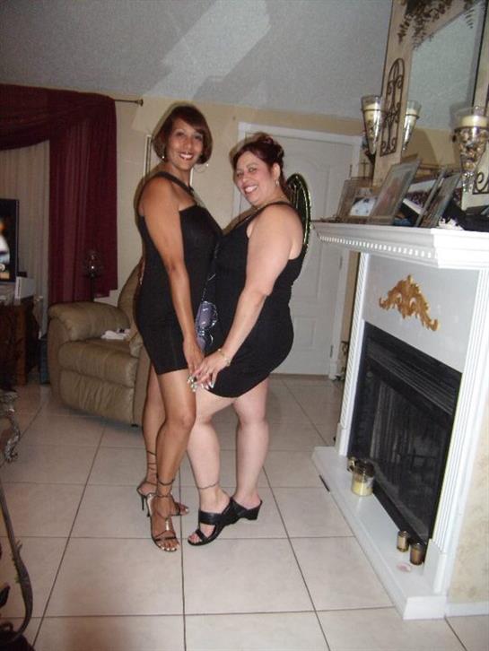 Two friends smile brightly while posing in fashionable black dresses at a home setting.