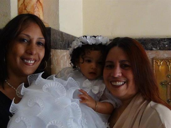 Two women smile joyfully as they hold a young girl in a white dress, celebrating family connections.