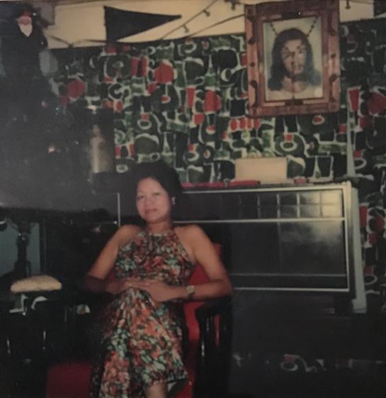 A woman relaxes in a chair wearing a colorful floral dress, surrounded by vintage decor.