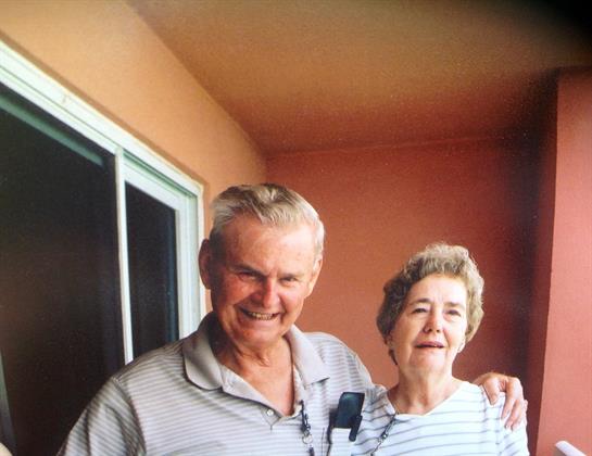 Two elderly people smile warmly while standing together on a balcony enjoying a sunny day.
